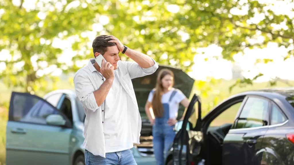 a man talking on a phone next to a car accident with a woman in the background