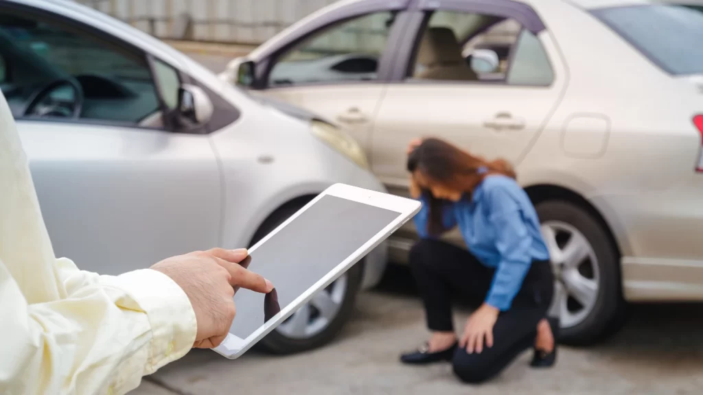a hand holding a tablet with a woman sitting on the ground next to a car accident